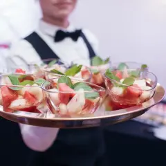 Taca z owocowymi deserami w szklanych miseczkach, podawana przez kelnera w białej koszuli i muszce.