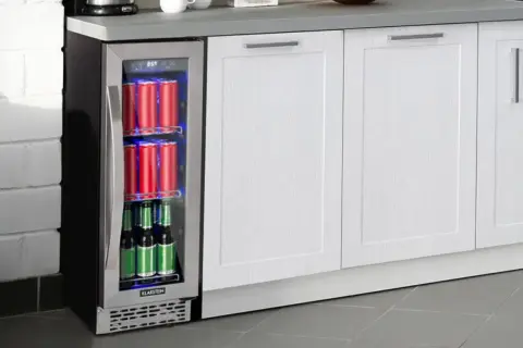 Szklana lodówka na napoje z czerwonymi puszkami i zielonymi butelkami, wbudowana w białą zabudowę kuchenną.