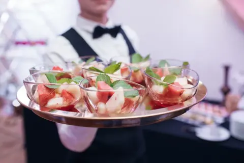 Taca z owocowymi deserami w szklanych miseczkach, podawana przez kelnera w białej koszuli i muszce.