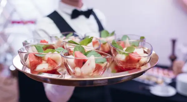 Taca z owocowymi deserami w szklanych miseczkach, podawana przez kelnera w białej koszuli i muszce.