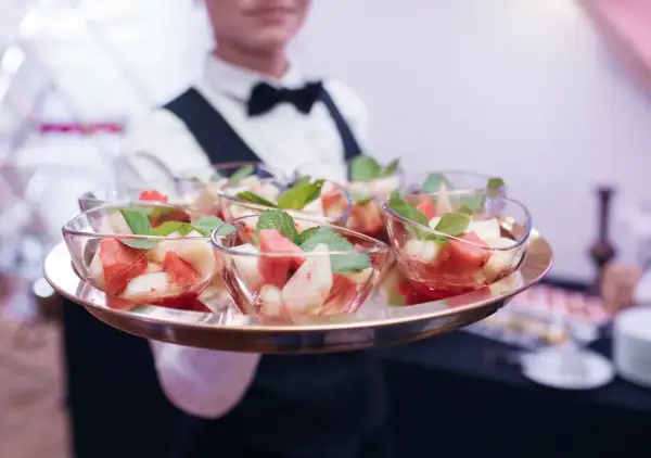 Taca z owocowymi deserami w szklanych miseczkach, podawana przez kelnera w białej koszuli i muszce.